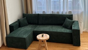 Een woonkamer met een groene hoekbank, een houten kruk en een groot raam met dunne gordijnen.