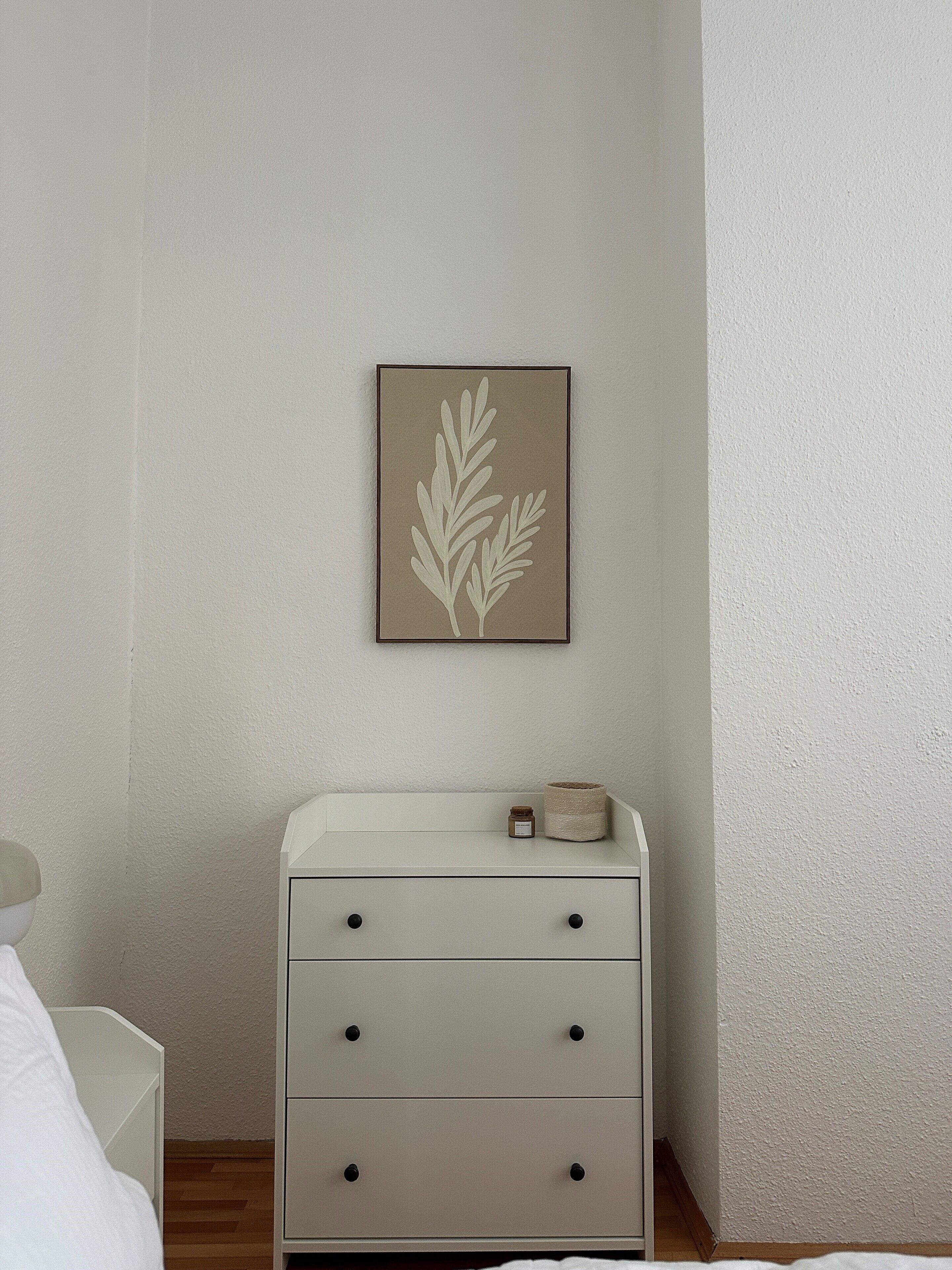 A bedroom with a white dresser, a framed leaf artwork on the wall, and a small jar on the dresser.
