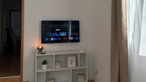 Een woonkamer met een flatscreen televisie aan de muur, een witte boekenkast eronder, en een houten vloer.