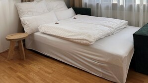 Een enkel bed met witte beddengoed, een houten kruk, een donkergroene bank en dunne gordijnen voor het raam.