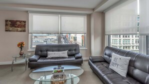 A modern living room with two leather sofas, a glass coffee table, and a view of a cityscape through large windows.