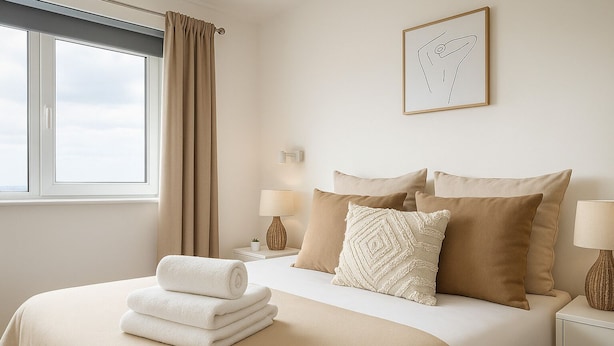 Una cama bien hecha con una colcha color beige, dos almohadas y una toalla enrollada. Se ve una ventana con cortinas color beige, una mesita de noche con una lámpara y un cuadro colgado en la pared.