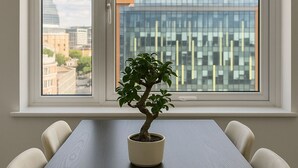 Una mesa de comedor con un bonsái en maceta, cuatro sillas y vistas a un edificio moderno a través de un amplio ventanal.