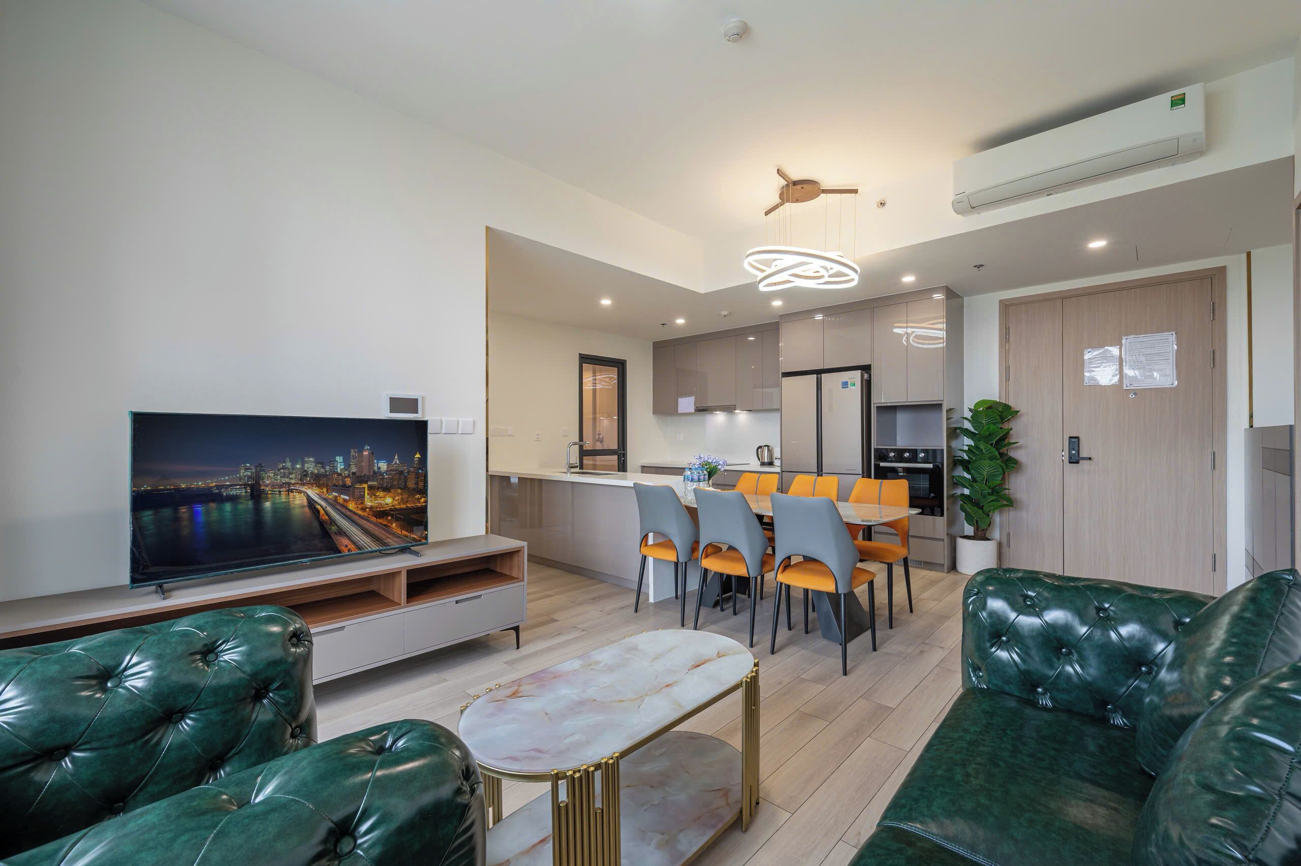A modern living room with a TV, green leather sofas, a dining area with a table and chairs, and a kitchen in the background.