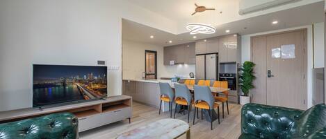 A modern living room with a TV, green leather sofas, a dining area with a table and chairs, and a kitchen in the background.