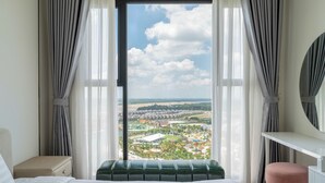 A hotel room with a large window, a bed, a green upholstered bench, a pink ottoman, and a white dresser with a mirror. - Studio - GH5 - Vinhomes Grand Park (Ho Chi Minh City)