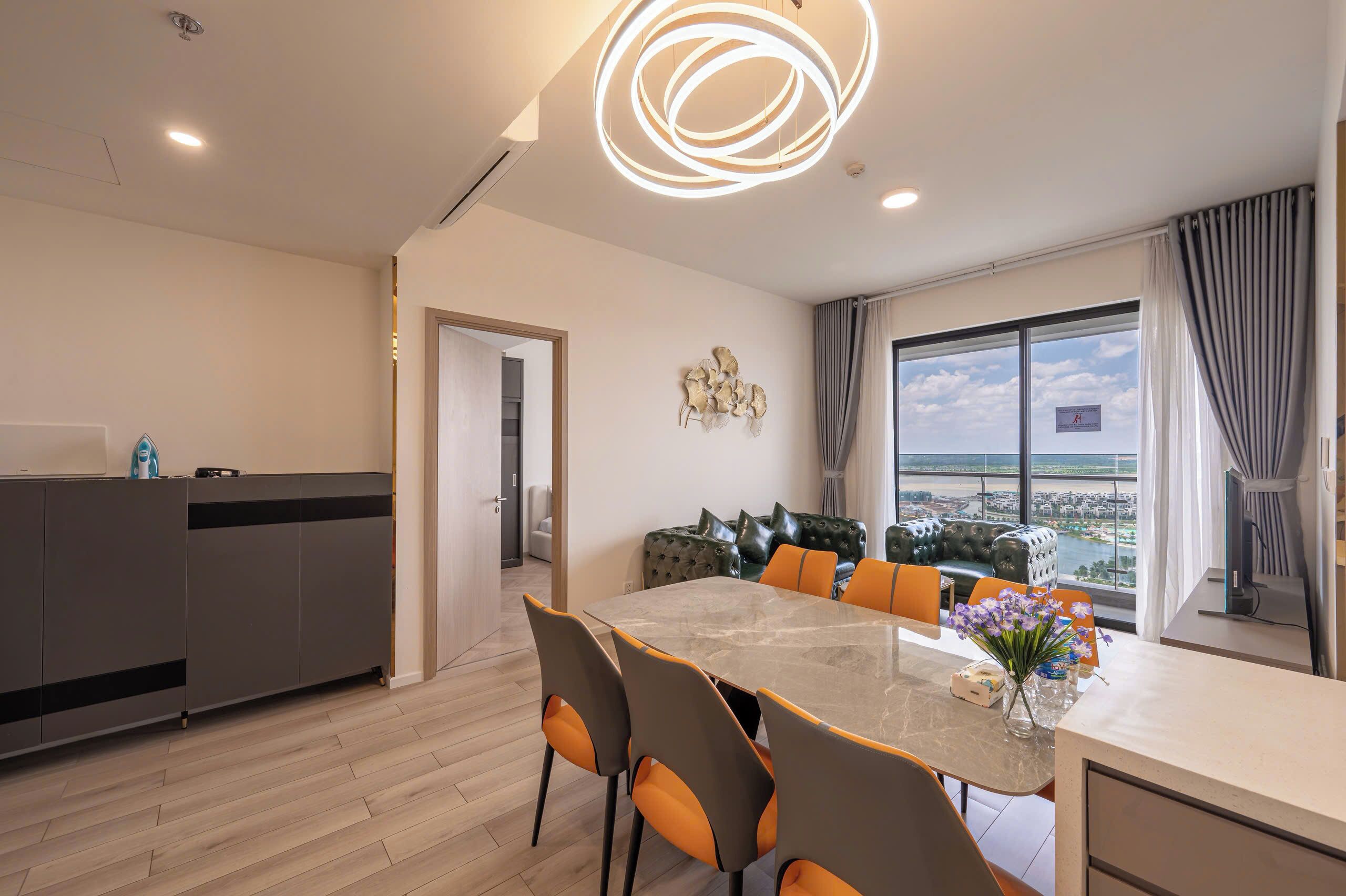 A modern dining area with a glass table and orange chairs, a grey sofa with decorative cushions, and a view of a body of water through large windows.