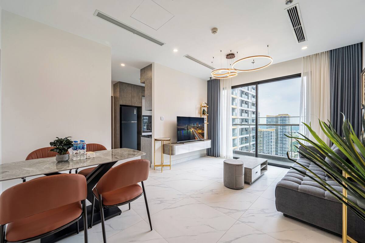 A modern living room with a large window, a dining area with a marble table, and a grey sofa.