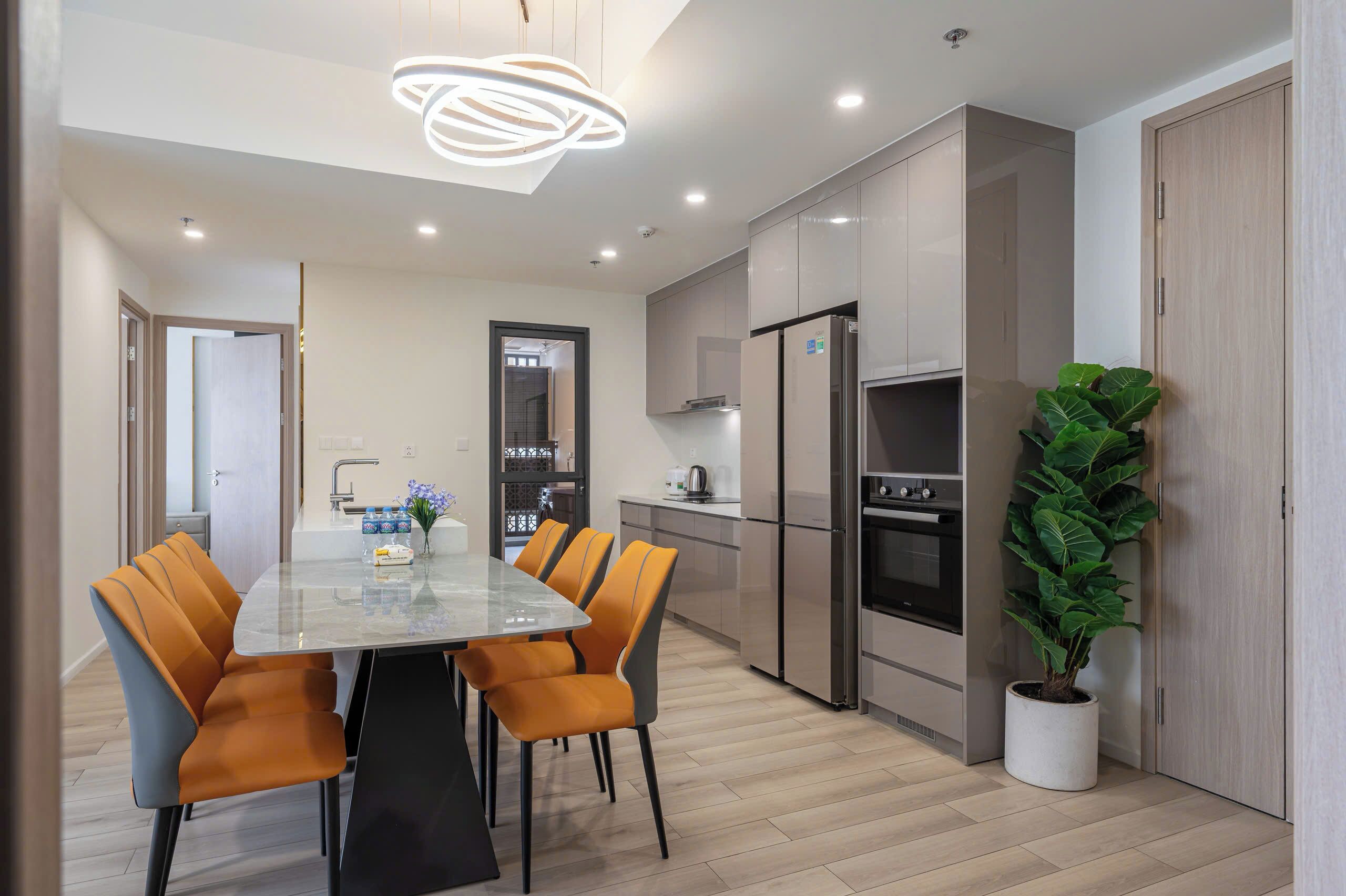 A modern kitchen with a dining area, featuring a large table, chairs, a refrigerator, oven, and a potted plant.