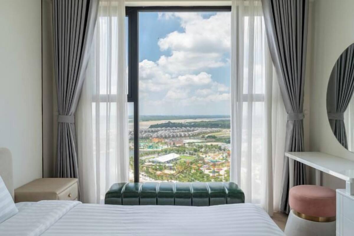 A hotel room with a large window, a bed, a green upholstered bench, a white bedside table, and a round mirror.
