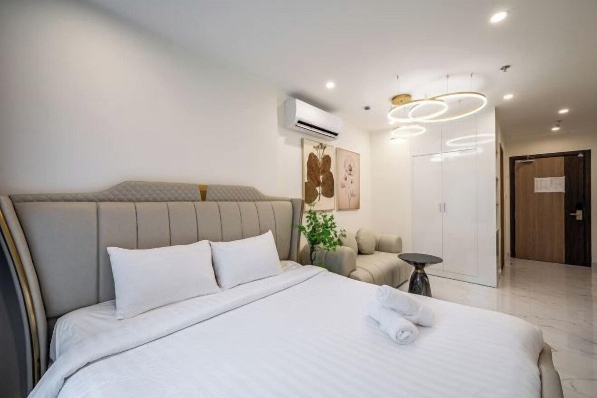 Quarto de hotel moderno com uma cama grande, mesa de cabeceira, ar condicionado, uma obra de arte na parede, um espaço de estar com uma cadeira e uma pequena mesa, e uma porta que dá para outro quarto.
