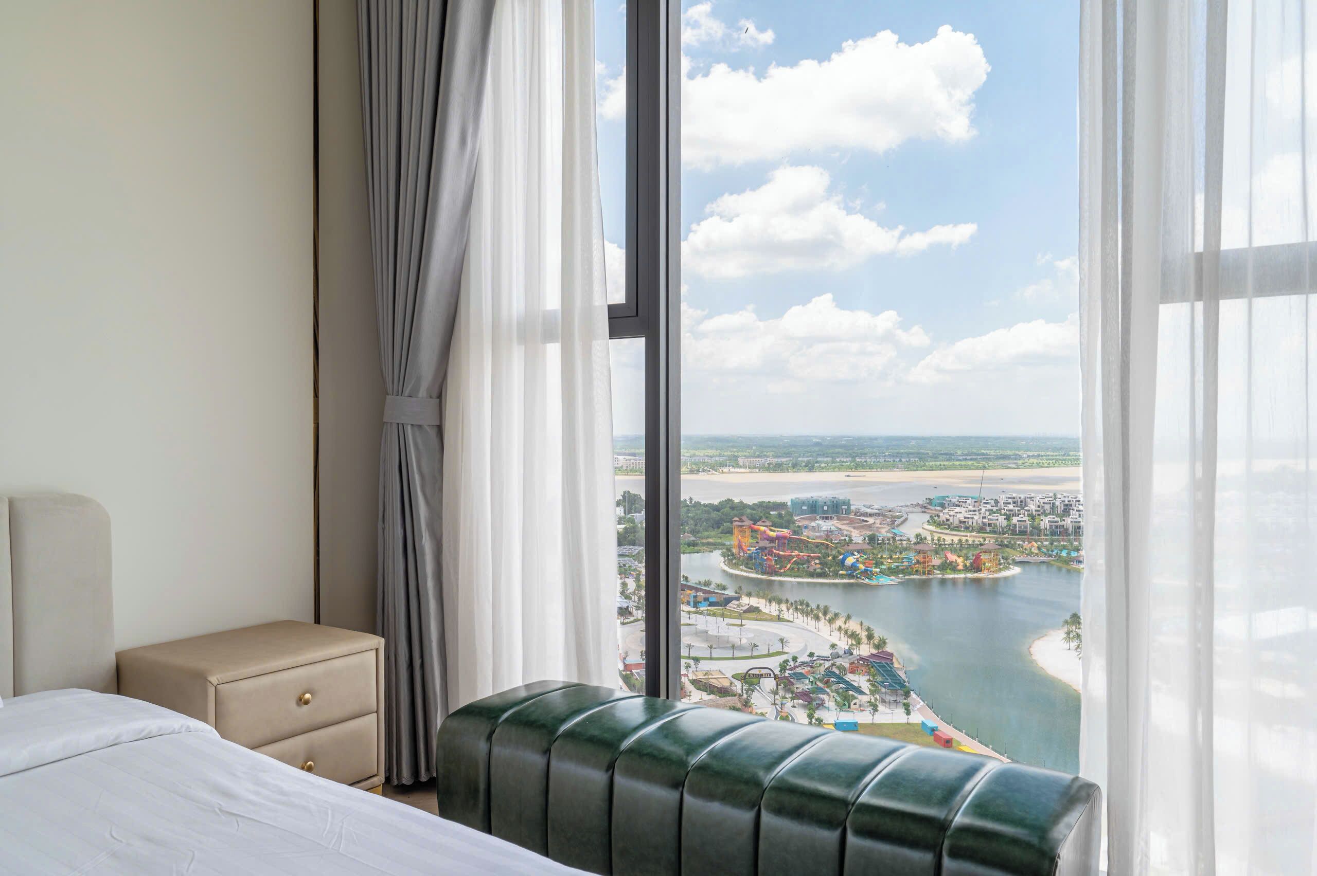 A hotel room with a bed, a small bedside table, a bench, and a large window offering a view of a coastal area with buildings and a body of water.