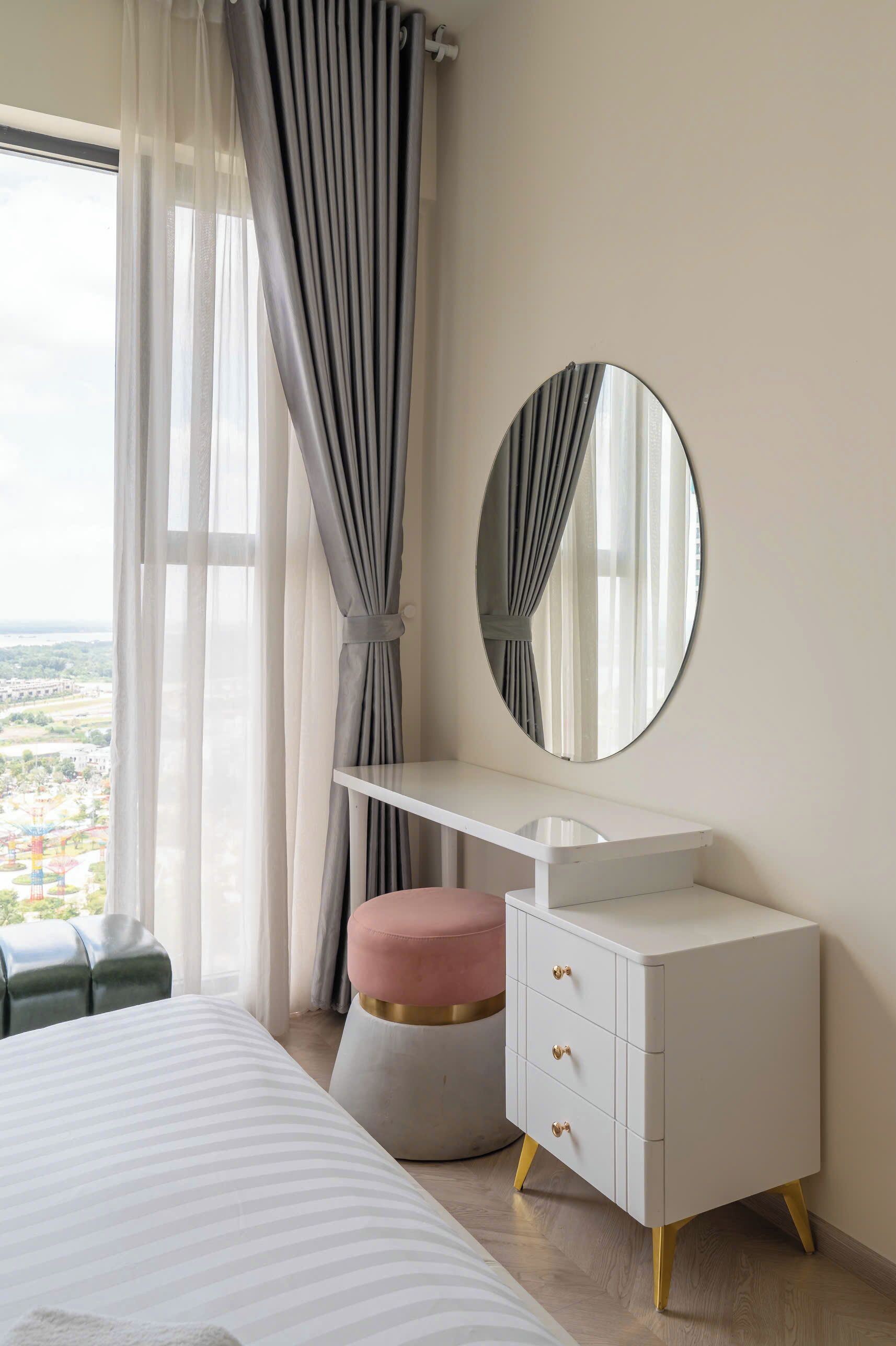 A bedroom with a bed, a white dresser with drawers, a round mirror, a pink ottoman, and a window with curtains.
