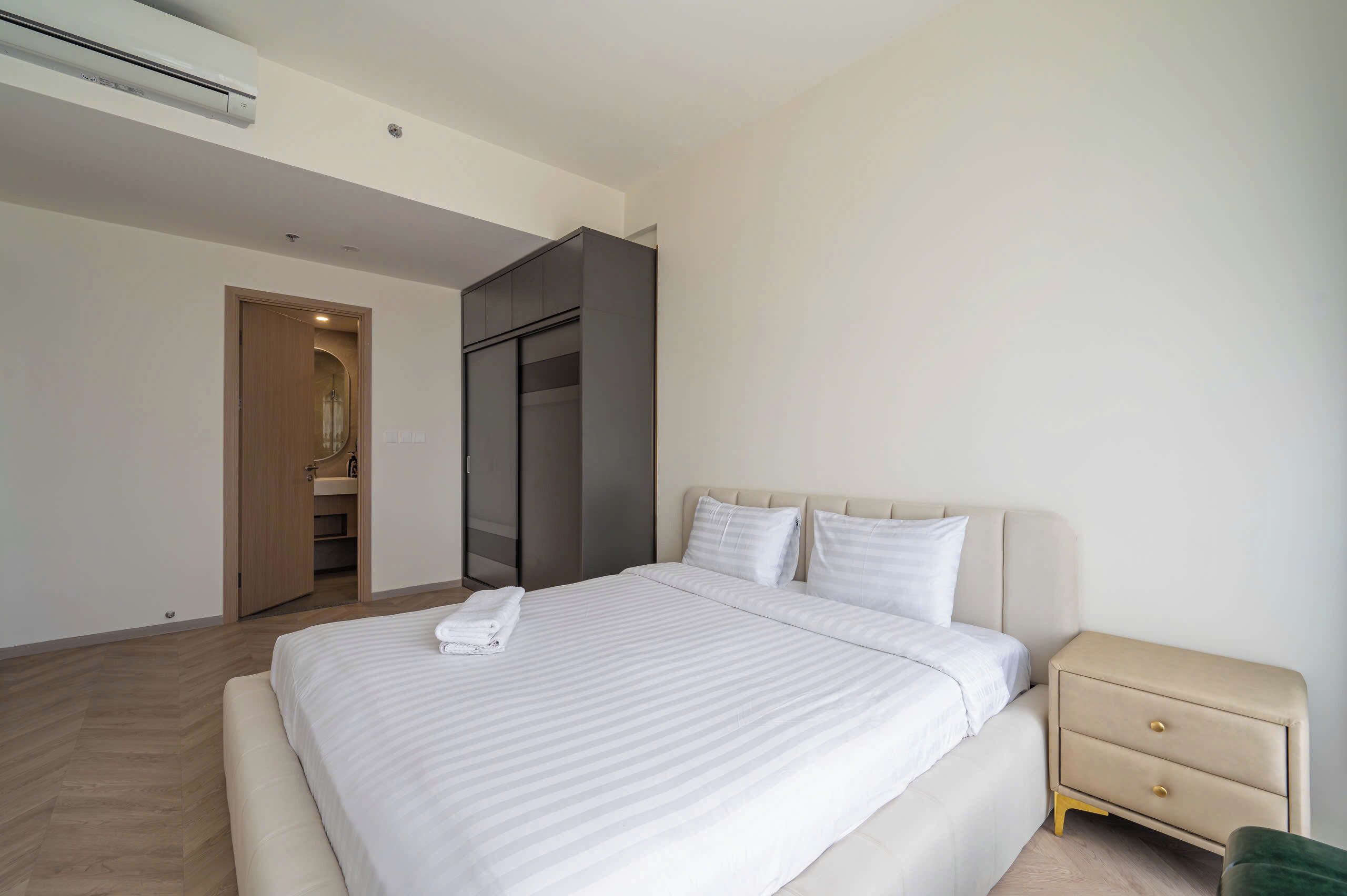 A modern hotel room with a large bed, a bedside table, a wardrobe, and a bathroom visible through an open door.