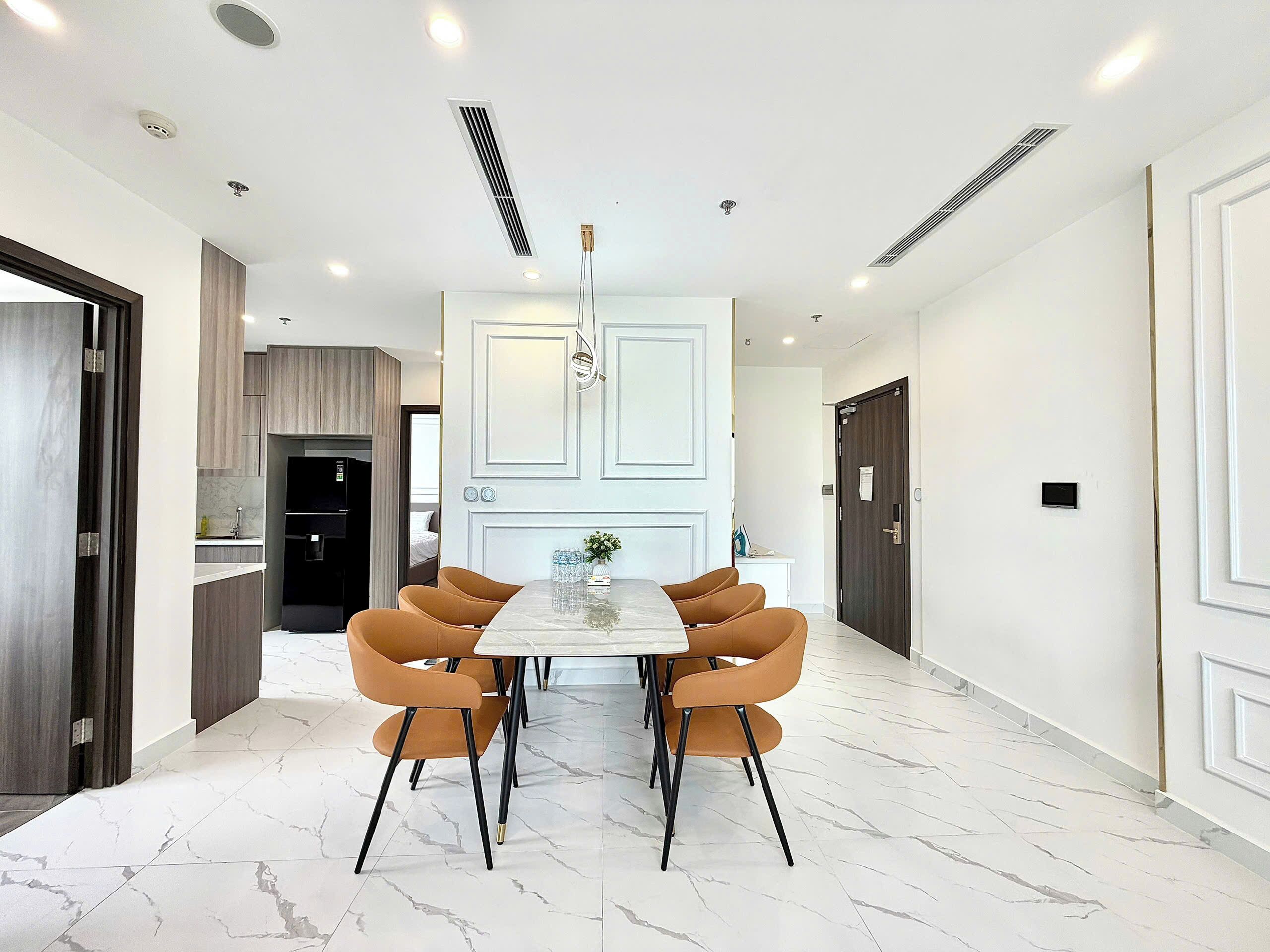 A modern hotel room with a marble floor, a dining area with a white table and brown chairs, a built-in kitchen, and a large wardrobe.
