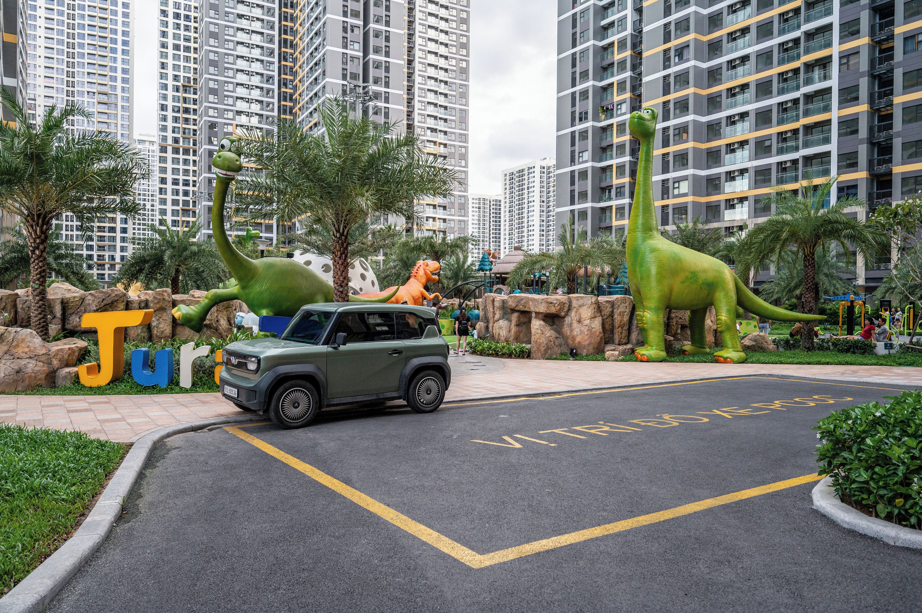 Un espace de stationnement avec un SUV vert, des statues de dinosaures et une pancarte indiquant « Jurassic Park ».