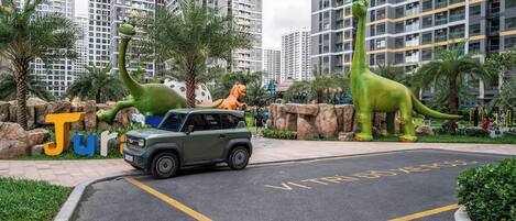 Un espace de stationnement avec un SUV vert, des statues de dinosaures et une pancarte indiquant « Jurassic Park ».