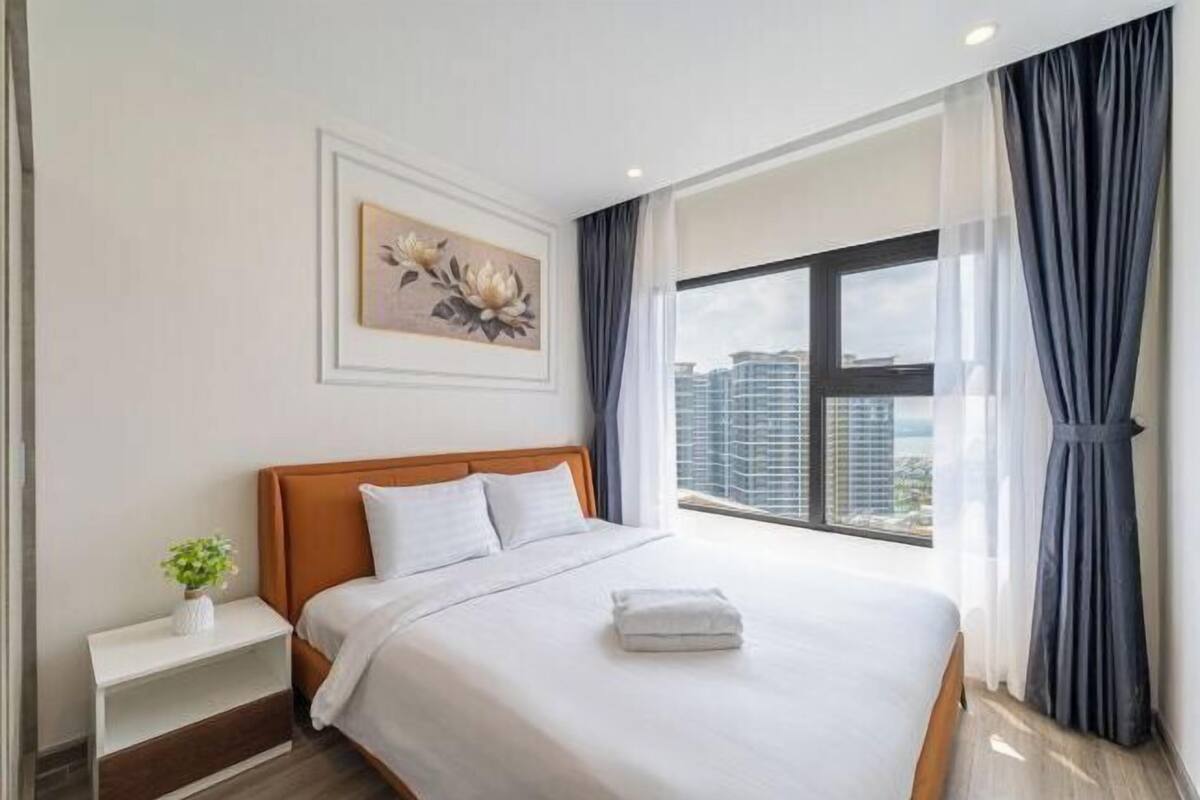 A modern hotel room with a large bed, a nightstand, a painting, and a view of the city.