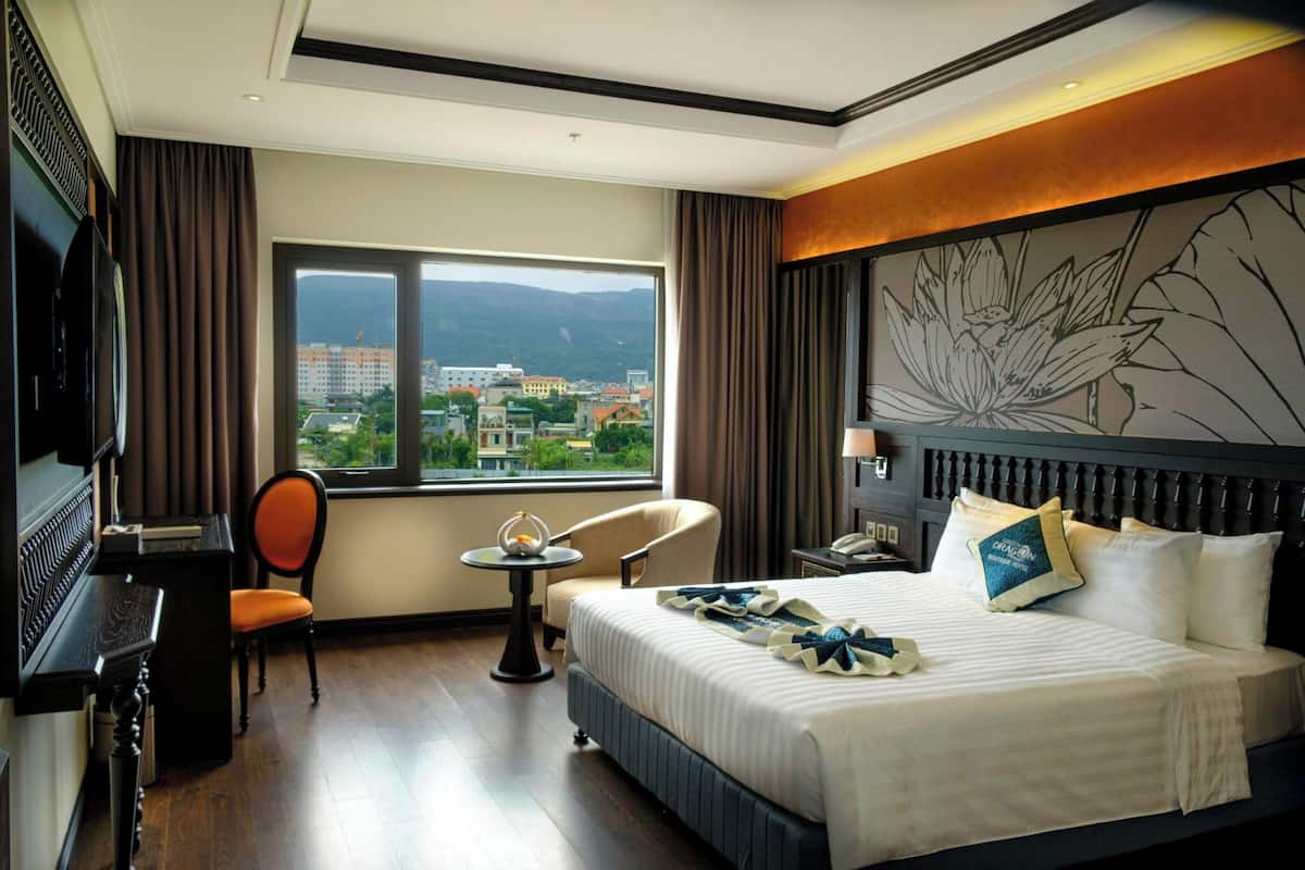 Habitación de hotel con una cama grande, un escritorio con televisor, una silla y vistas a un paisaje urbano a través de un ventanal amplio.