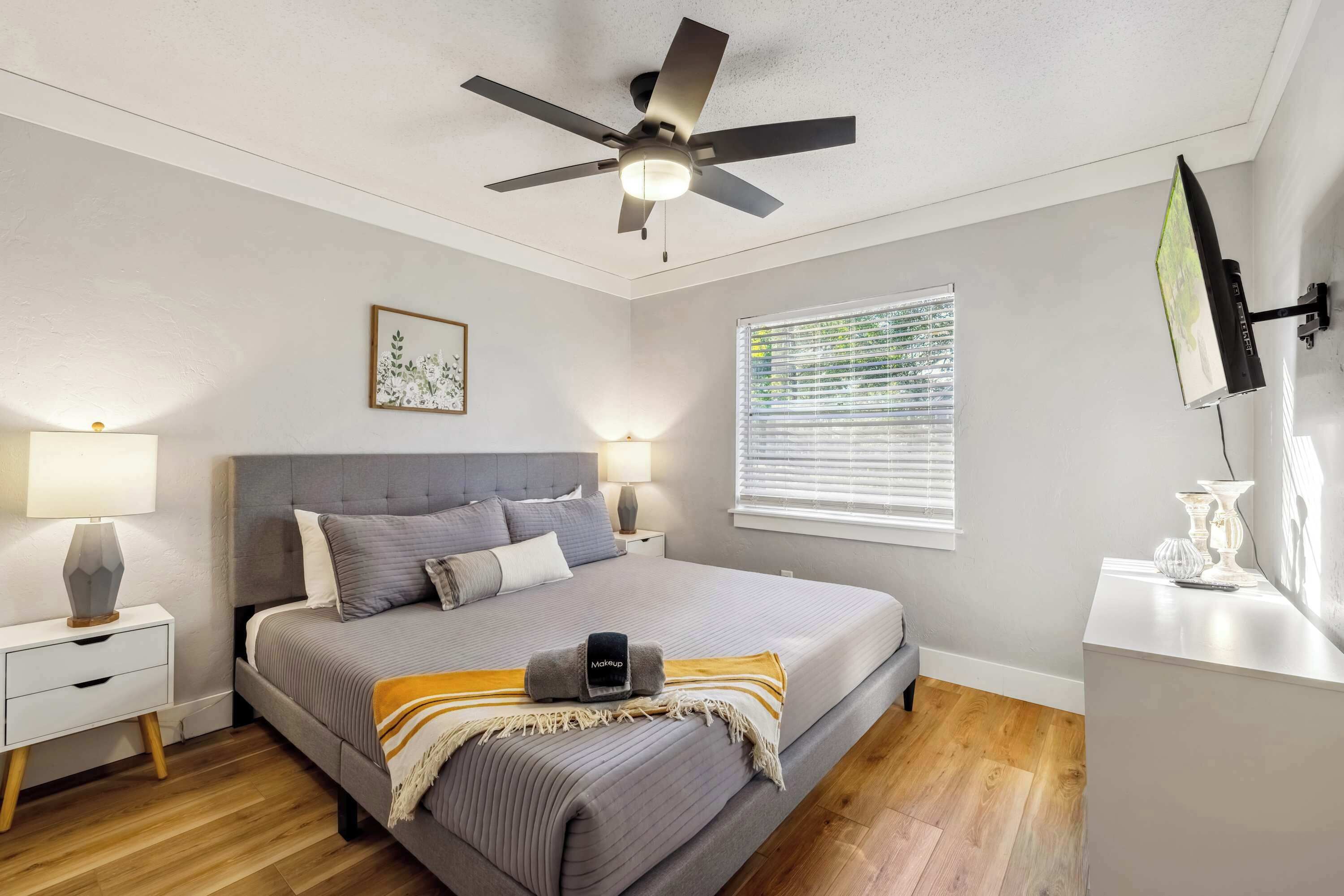 A bedroom with a bed, bedside lamps, a ceiling fan, a window with blinds, and a wall-mounted TV.