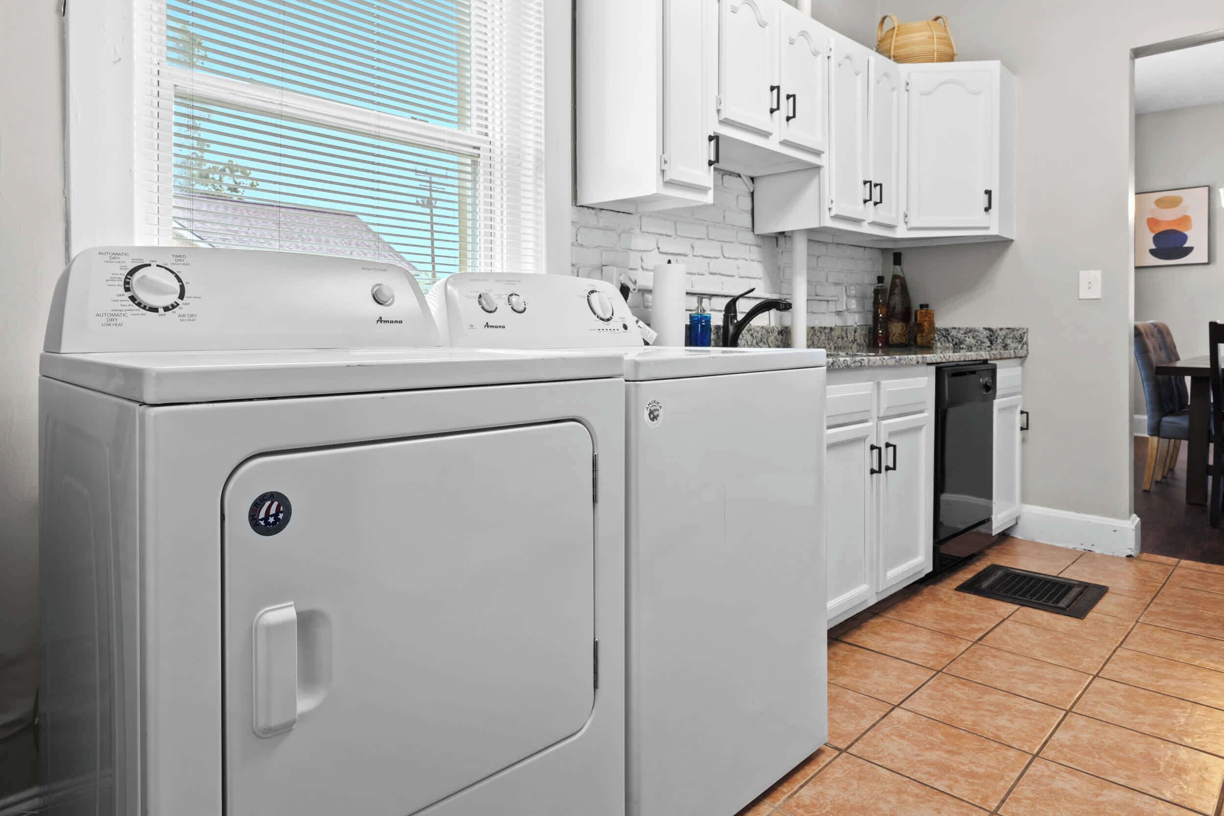A laundry room with two front-loading washing machines, a countertop with a sink, cabinets, and a window with blinds.