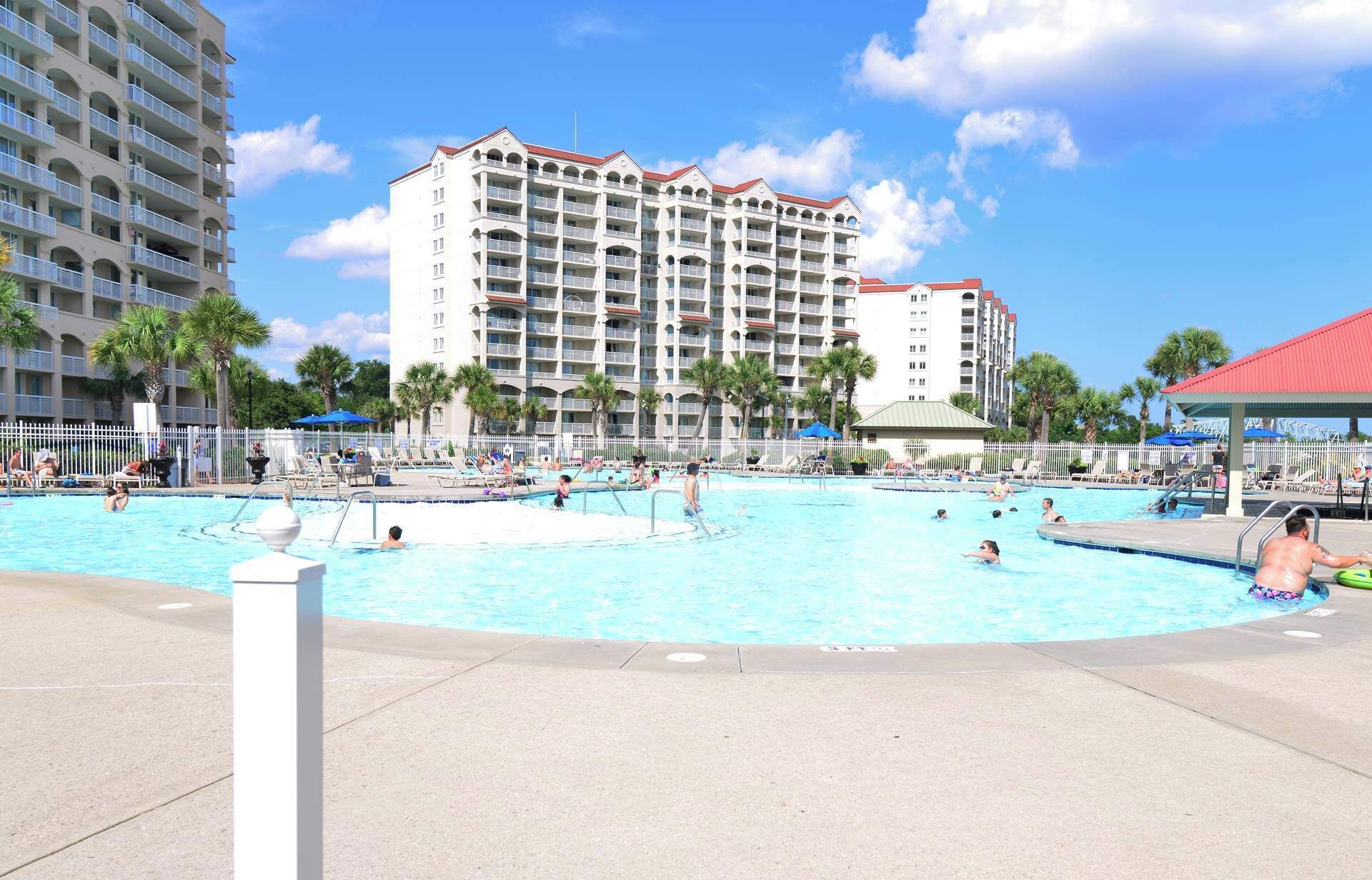 A large outdoor swimming pool surrounded by palm trees and a hotel building with multiple balconies.
