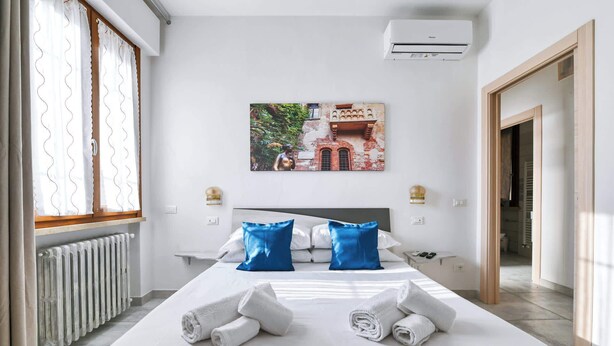 A hotel room with a bed, white linens, two blue pillows, a painting of a building, an air conditioner, and a radiator. - The Red House - Verona Holiday & Business (Verona)