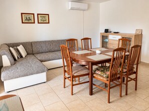 Een woonkamer met een grijze bank, een houten eettafel en stoelen, een witte airconditioner en een klein kastje met een magnetron.