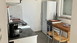 Kompakti keittiö, jossa on kaasuliesi, valkoinen jääkaappi, pieni ruokapöytä ja kaksi rottinkituolia sekä ikkuna, jossa on sälekaihtimet.