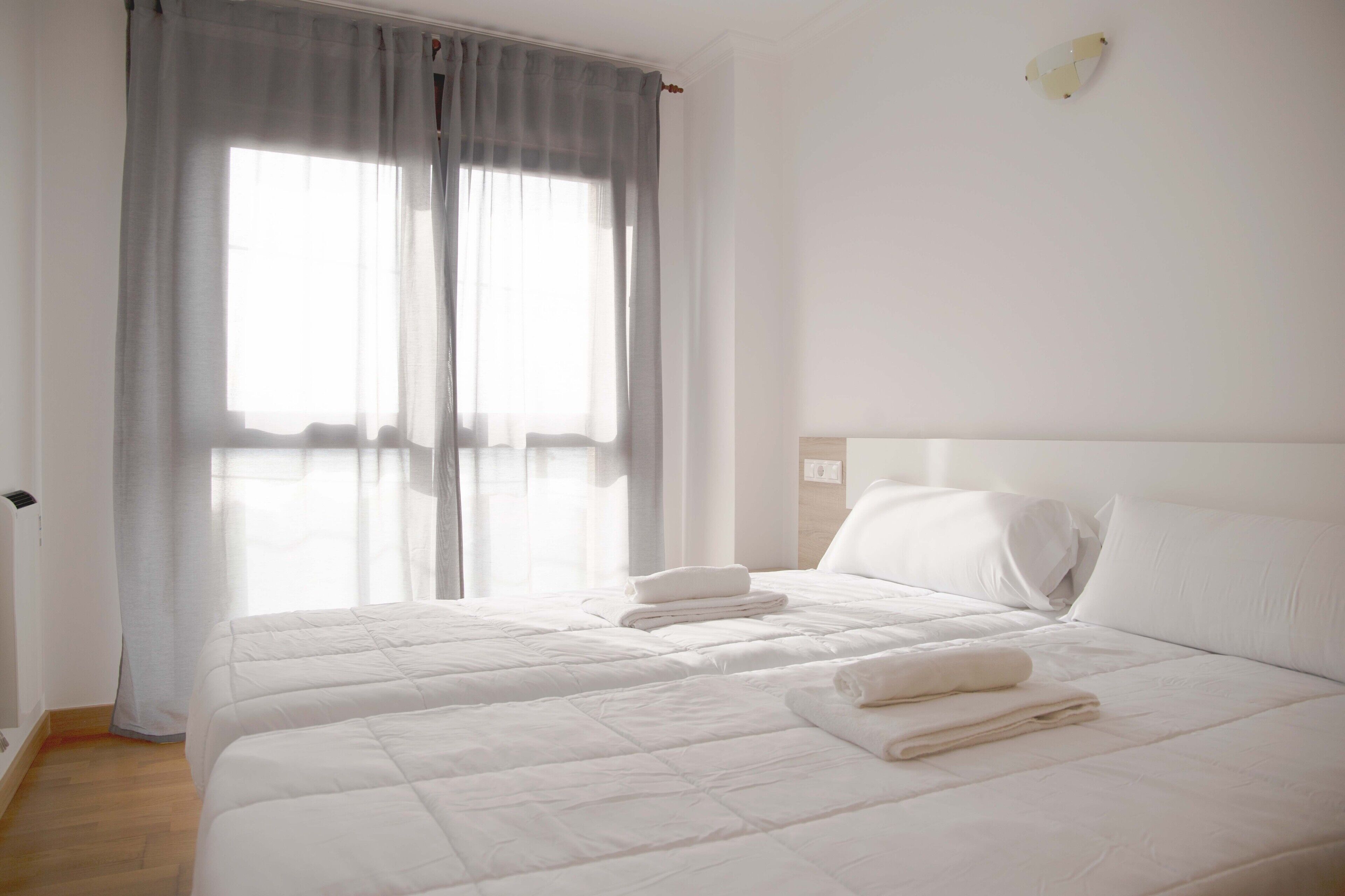 Quarto de hotel com duas camas, uma janela grande com cortinas e cabeceira branca.