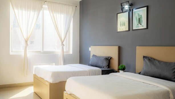A hotel room with two beds, a window with curtains, a bedside table with a plant, and two framed pictures on the wall. - Housr Sector 69 Gurgaon Near Sohna Road (Gurugram)