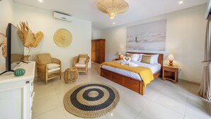 Un dormitorio amplio con una cama grande, dos mesitas de noche, un armario de madera, una silla de mimbre, un tapete redondo y un aire acondicionado instalado en la pared.