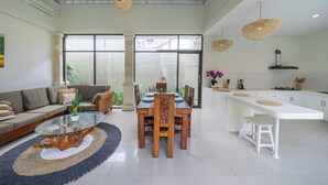 Amplia sala de estar con una mesa de centro de vidrio, un sofá y una mesa de comedor con sillas. Se observa una zona de cocina con alacenas blancas y una encimera.