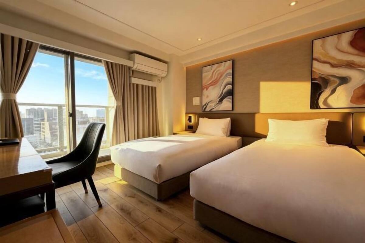 Habitación de hotel con dos camas, un escritorio, una silla y ventanas grandes con cortinas.