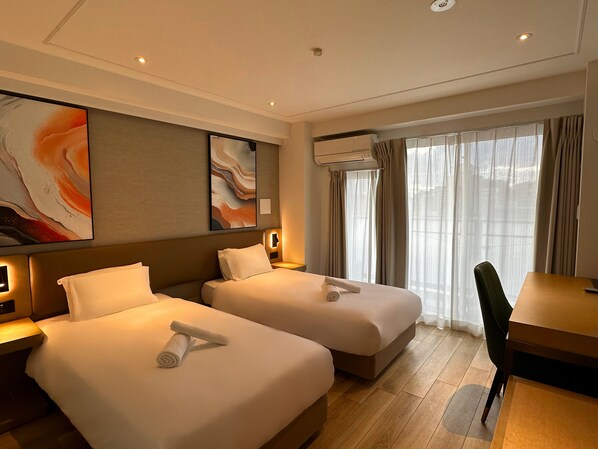 Ett hotellrum med två sängar, ett skrivbord och en stol. Det finns två väggmonterade luftkonditioneringsapparater och ett stort fönster med gardiner.