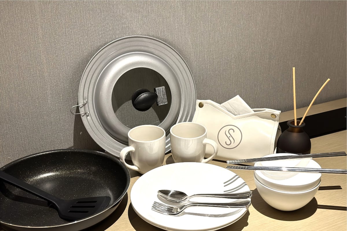 Una disposición para el desayuno con una sartén, dos tazas blancas, un plato con cubiertos, un cuenco pequeño y un adorno con ramitas.