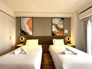 Ett hotellrum med två sängar, var och en med ett vitt överkast och en hoprullad handduk. På väggen ovanför sängarna hänger abstrakta målningar, och i taket finns en taklampa.