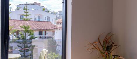 A room with a bed, a chair, a potted plant, and a view through a glass door.