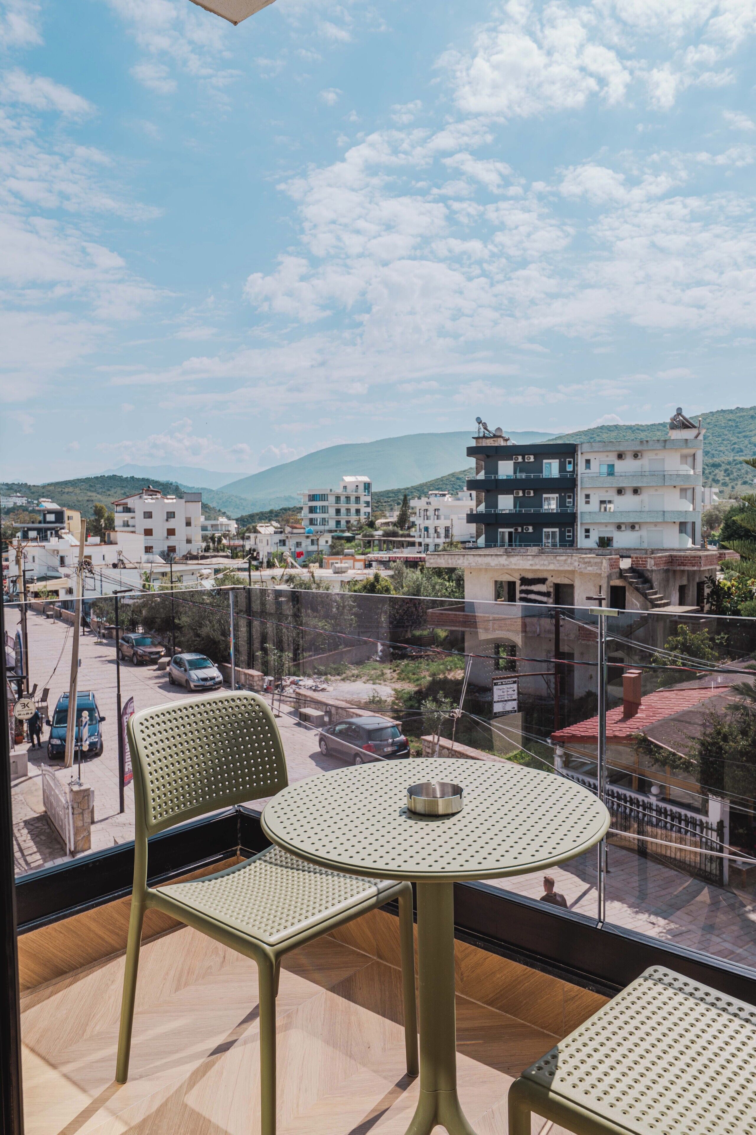 A balcony with two green chairs and a small table, overlooking a street with buildings and mountains in the background.