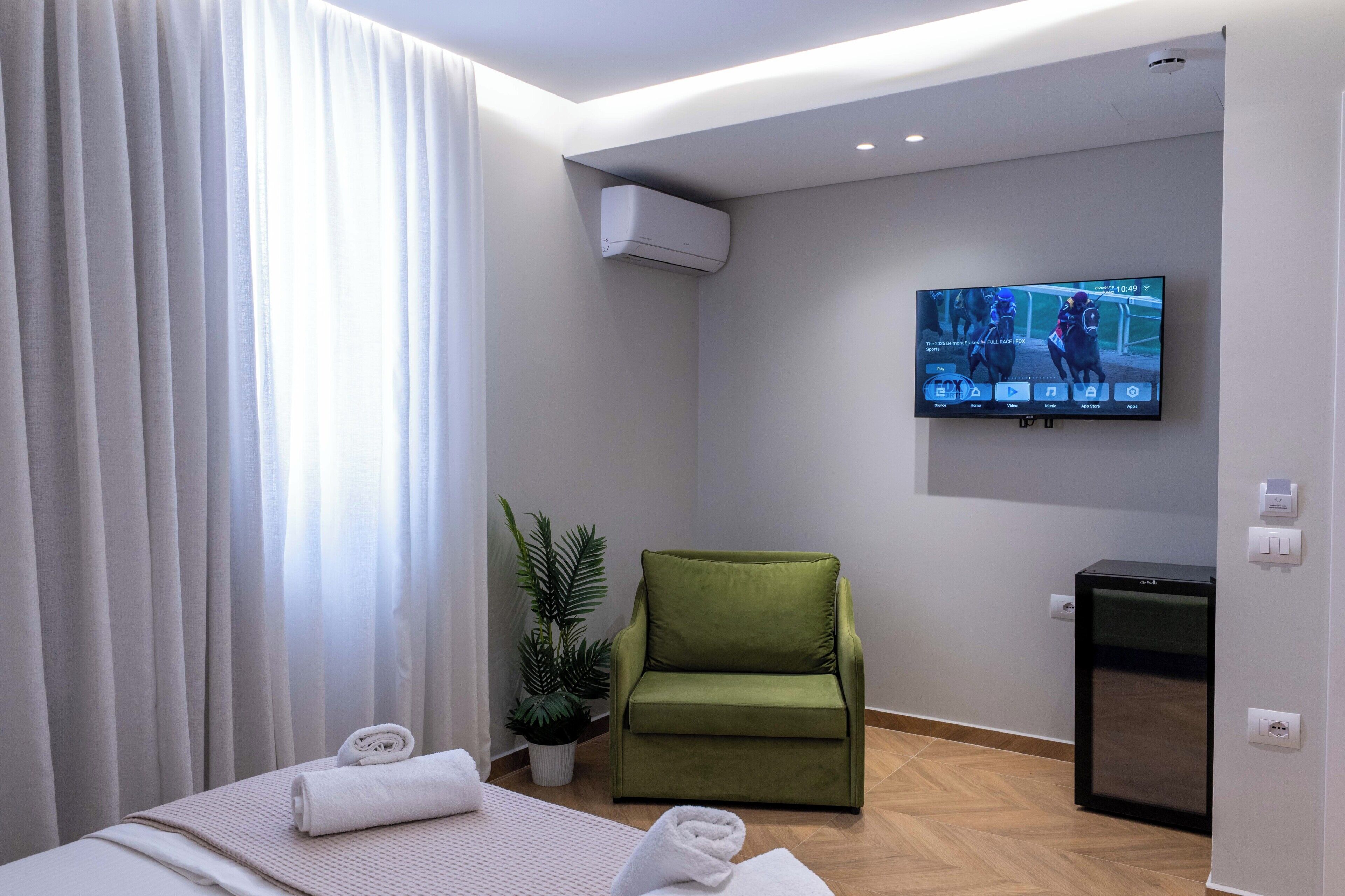 A hotel room with a bed, a green armchair, a television, a small refrigerator, and a potted plant.