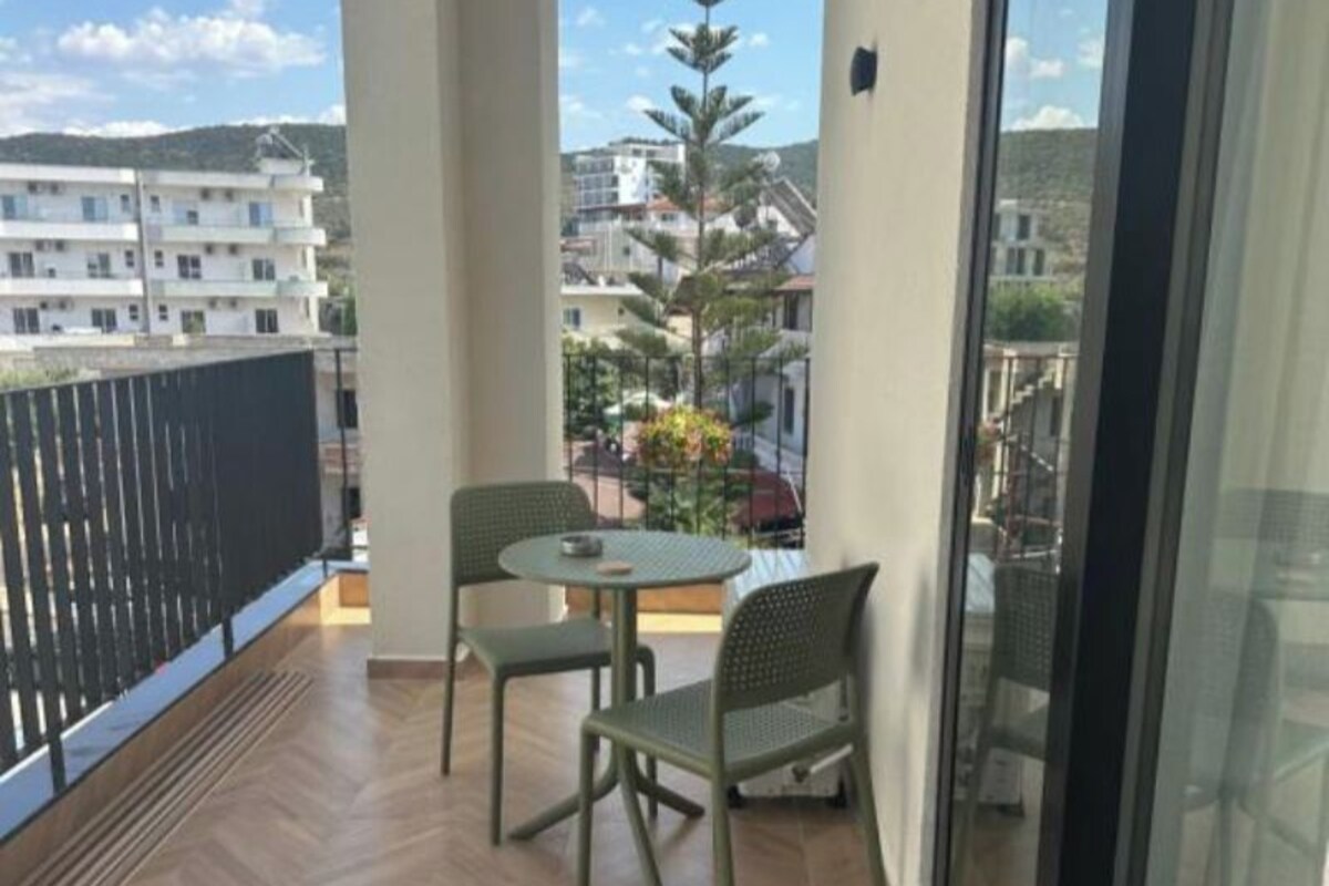 A balcony with two chairs and a table, wooden flooring, and a view of buildings and trees.