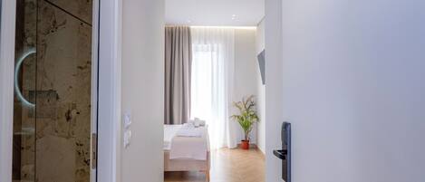 A hallway leading to a bedroom with a bed, a potted plant, and a glass shower area.