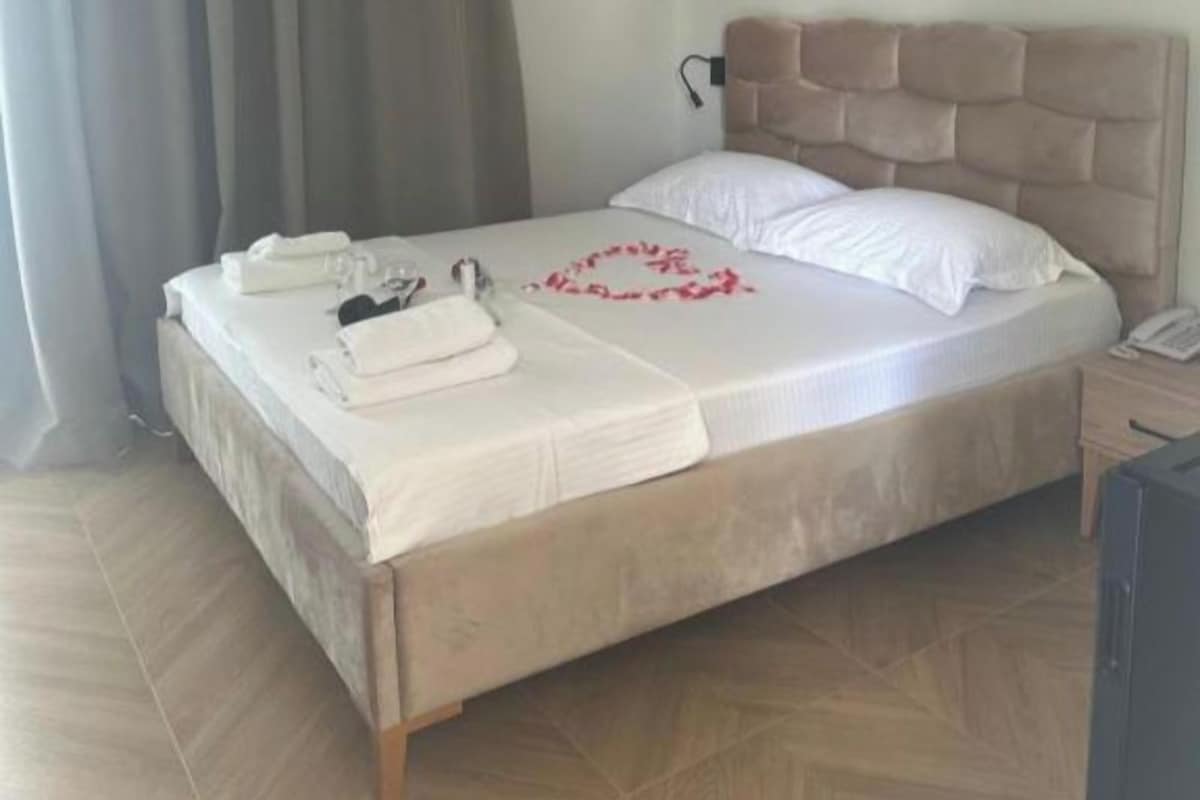 Cama bem arrumada com edredom branco, um coração decorativo vermelho e toalhas dobradas ao lado.
