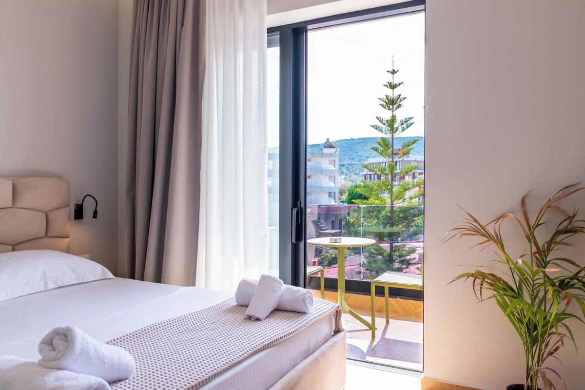A hotel room with a bed, a window with curtains, a small balcony with a table and chair, and a potted plant.