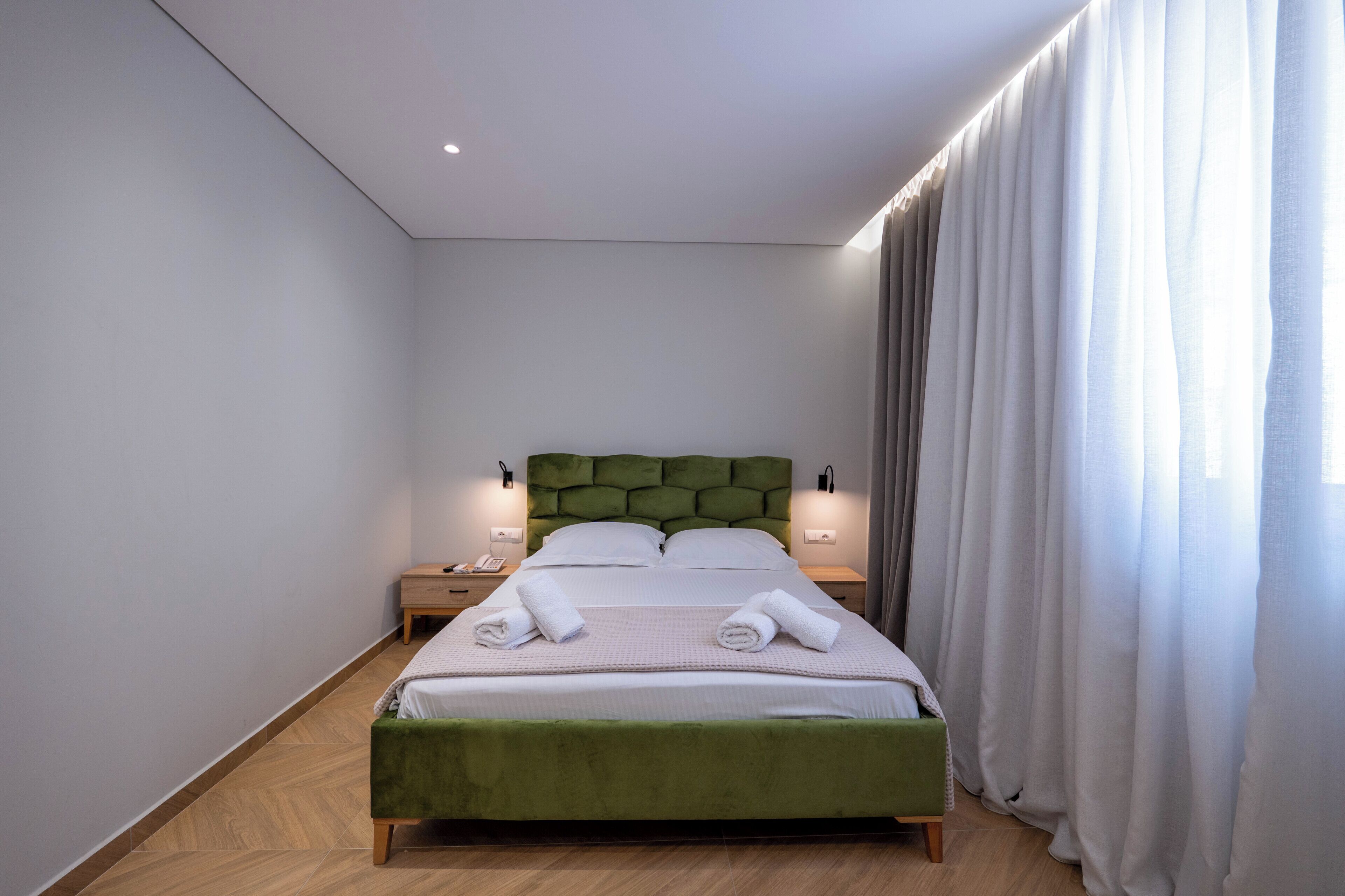 A neatly made bed with pillows and towels, a green upholstered headboard, bedside tables with lamps, and a window with curtains.