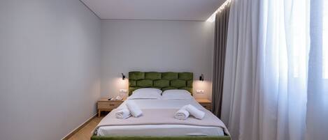 A neatly made bed with pillows and towels, a green upholstered headboard, bedside tables with lamps, and a window with curtains.