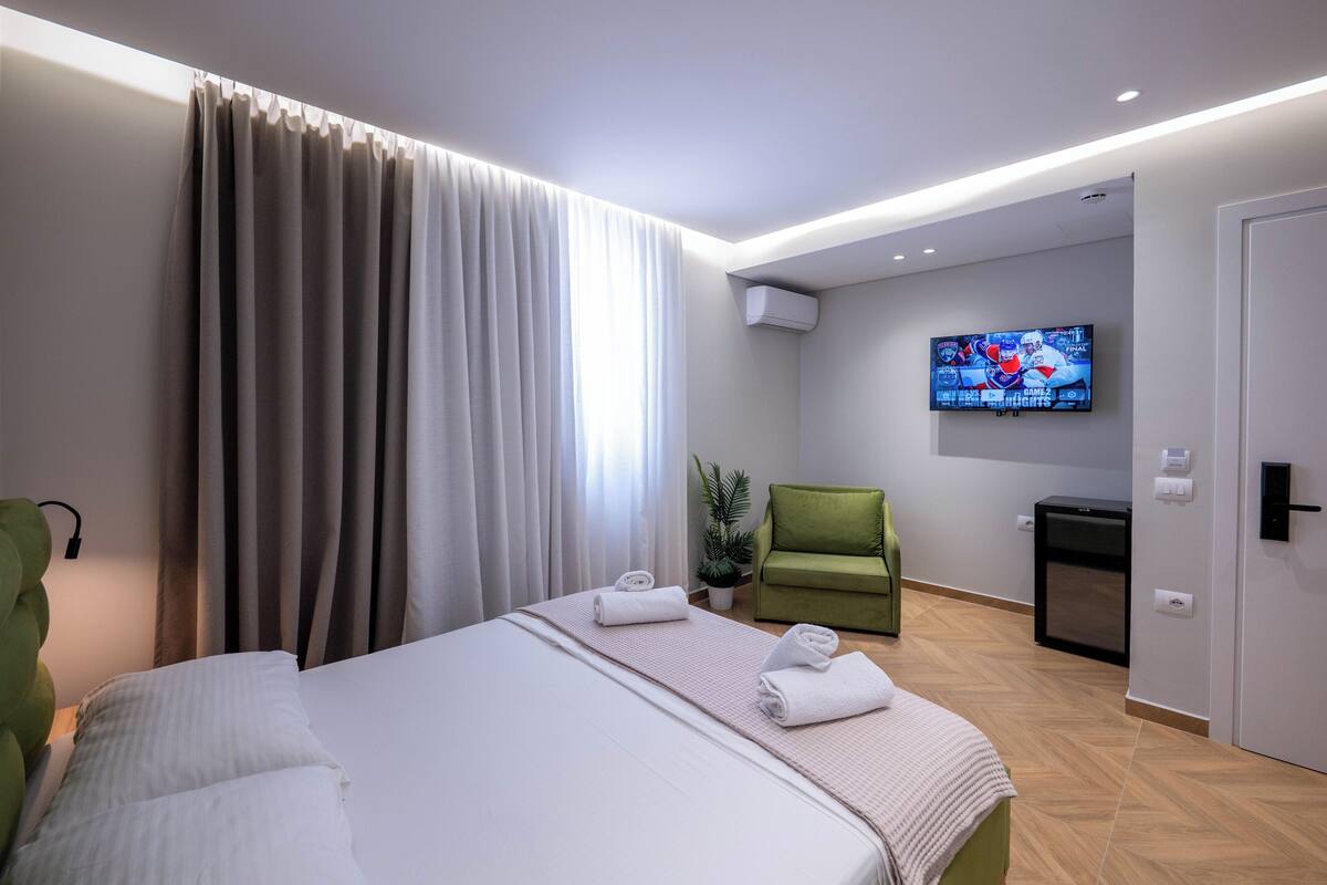 A hotel room with a bed, a TV mounted on the wall, a green chair, and a small table.