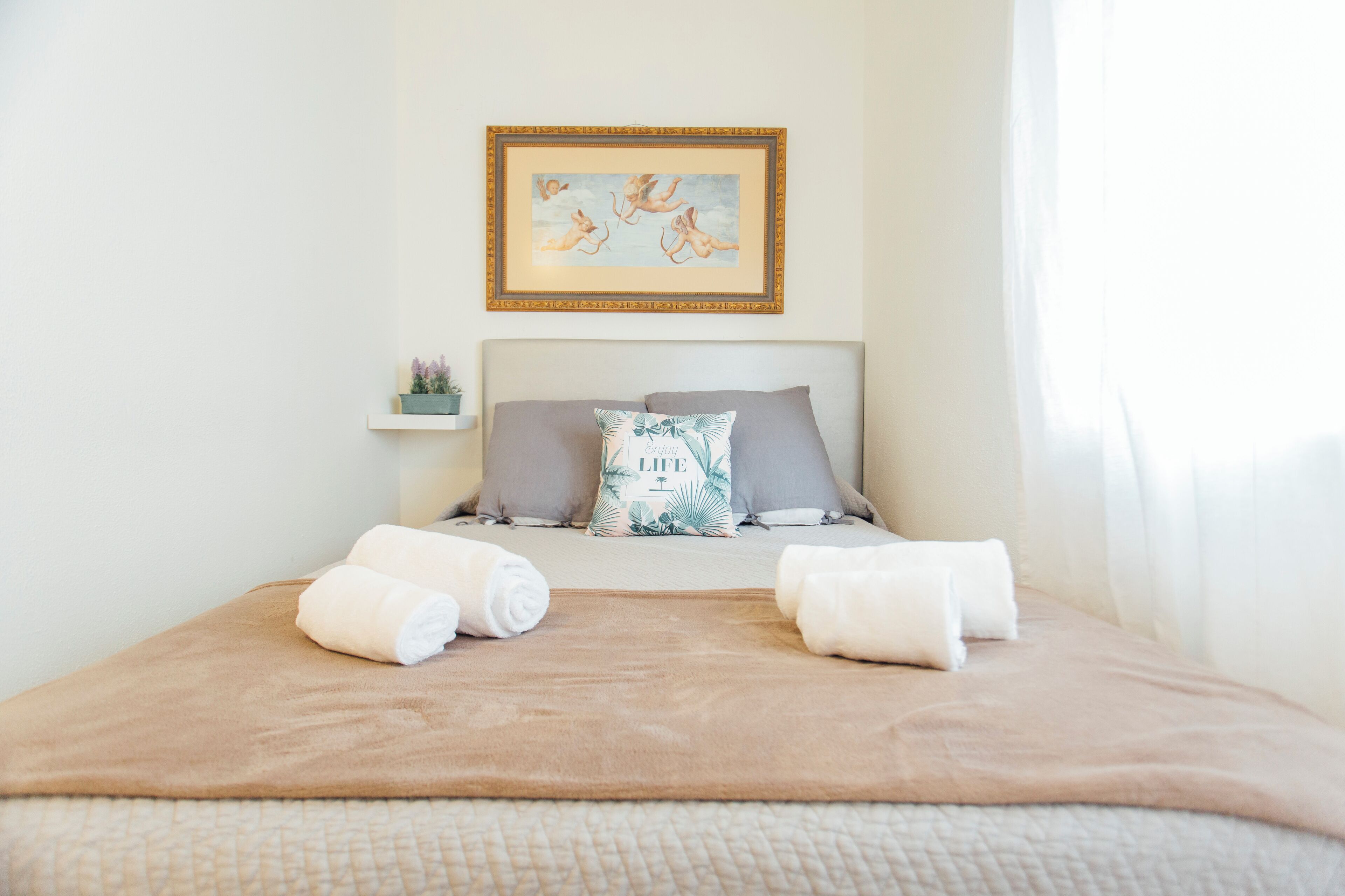 Un letto rifatto con cura, con una coperta e cuscini color beige, un quadro appeso sopra e un piccolo ripiano con una pianta.