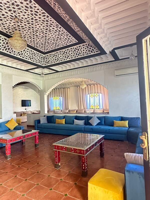 A spacious living room with a blue sofa, red and yellow cushions, a glass coffee table, and a decorative ceiling.