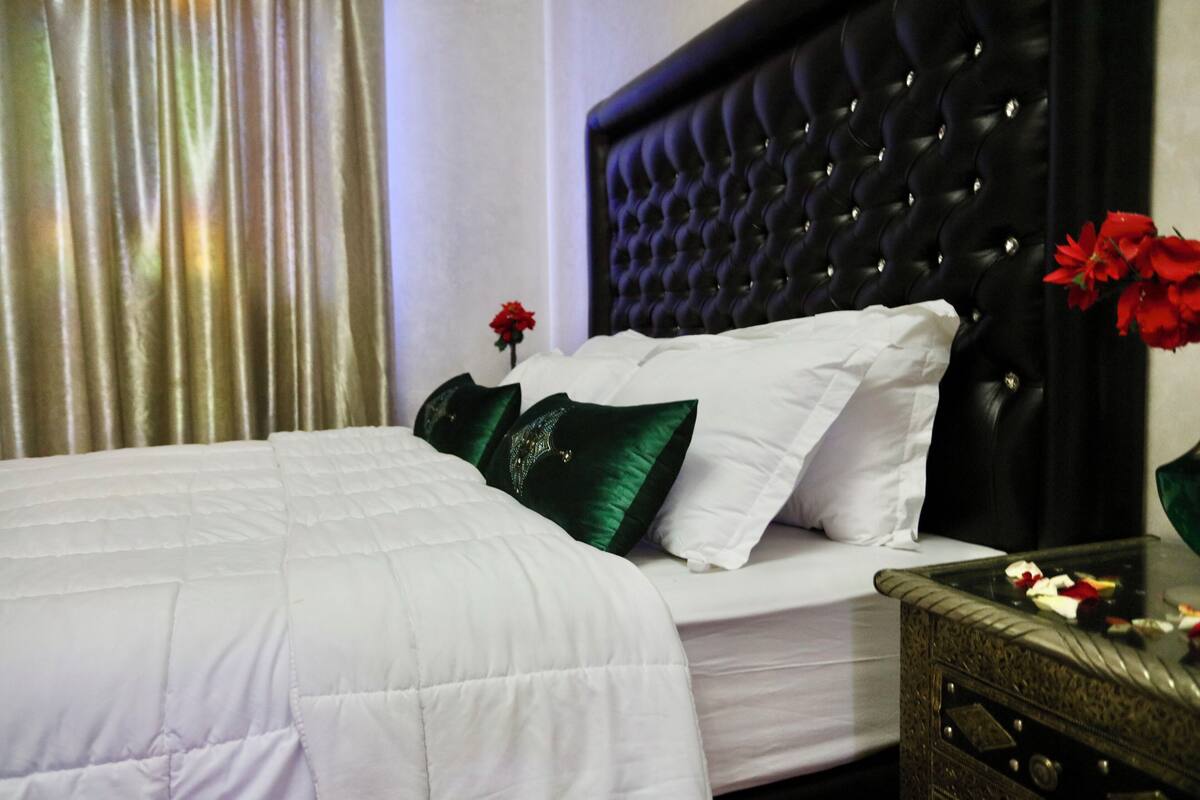 Una habitación de hotel con una cama grande, un cabecero capitonado y una mesita de noche con un jarrón de flores.